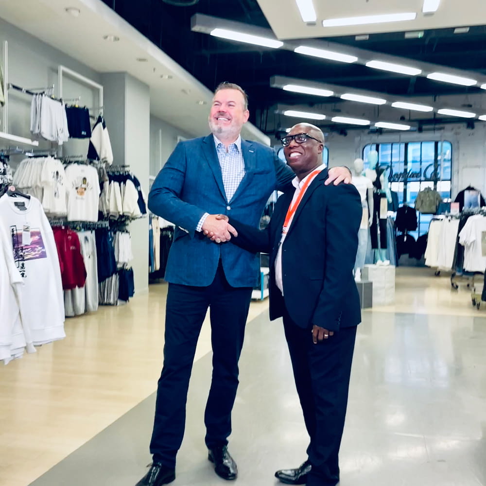 Two men in suits shake hands and smile inside a modern clothing shop. Shelves and rails of clothes are visible in the background under bright ceiling lights.