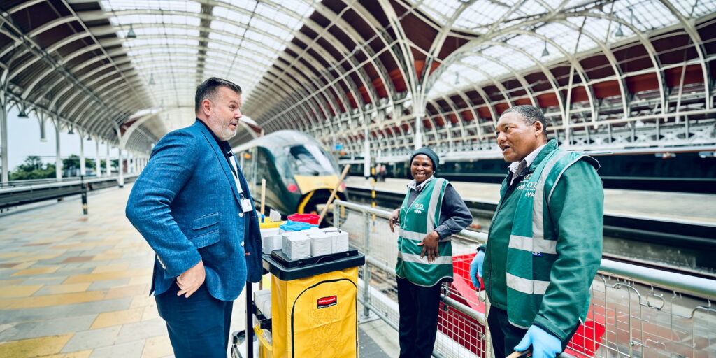 A man in a blue suit talks with two railway station workers in green vests beside a yellow cleaning trolley on a covered railway platform, with a train in the background.