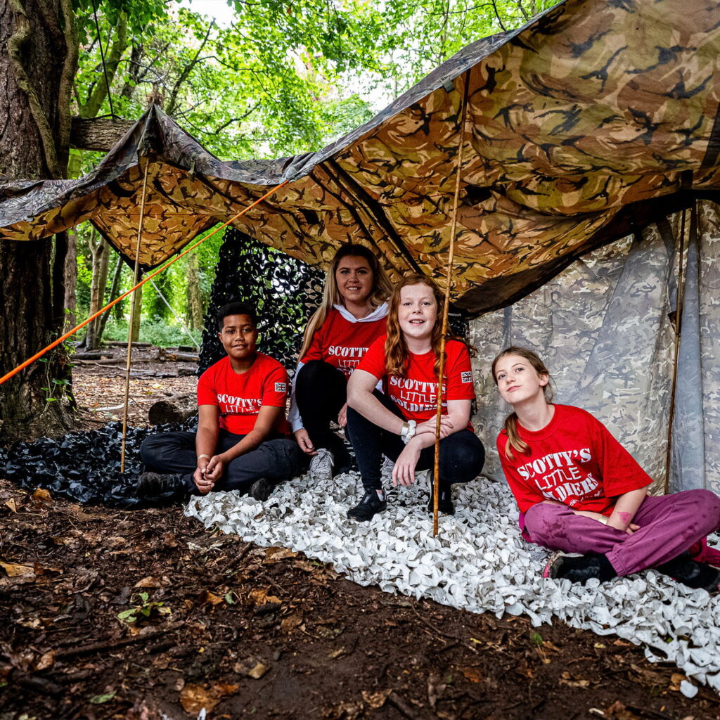 Four children in red Scotty's Little Soldiers shirts sit and kneel under a camouflage shelter in a wooded area, surrounded by trees and leaves. They look at the camera, smiling and relaxed.
