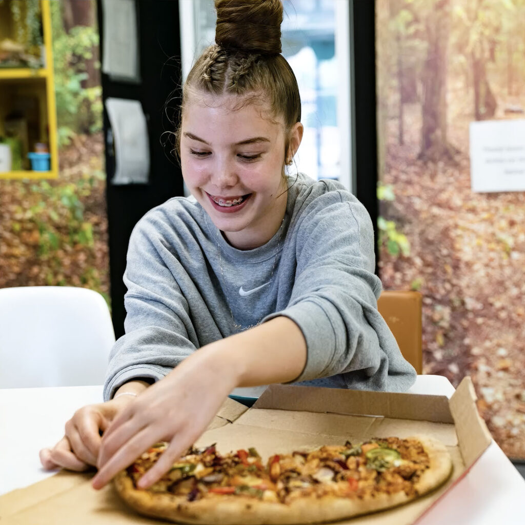 A smiling girl with braces reaches for a slice of pizza from a box while sitting at a table. She wears a grey sweatshirt and has her long hair styled in a high bun. The background features a forest-themed wall mural.