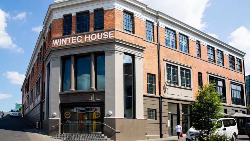 A large brick and concrete building with WINTEC HOUSE in bold letters near the roof; several large windows, a glass entrance, a tree, a white van, and a person walking on the pavement nearby.