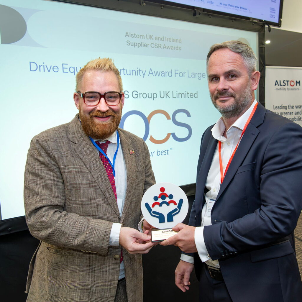 Two men in suits smiling and holding an award together. Behind them is a screen displaying Drive Equal Opportunity Award For Large, OCS Group UK Limited, and the Alstom UK and Ireland Supplier CSR Awards logo.
