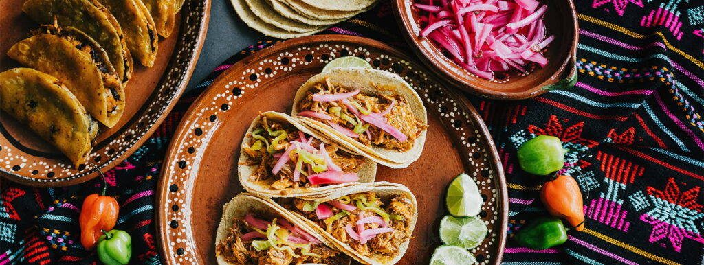 Three tacos with shredded meat, pickled onions, and limes on a decorated plate, surrounded by tortillas, more pickled onions, colourful cloth, and peppers.