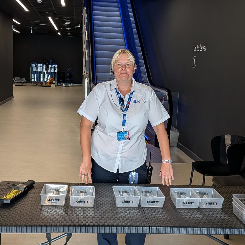 A security staff member stands behind a table with clear trays labelled GARRETT, near an escalator in a modern indoor area.
