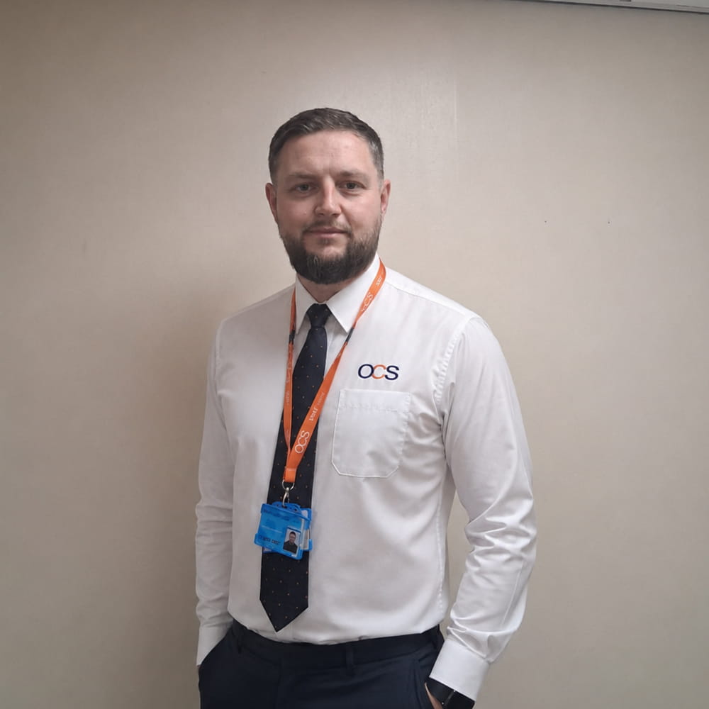 A man with a short beard stands against a plain wall, wearing a white shirt with an “OCS” logo, a black tie, and an orange lanyard with blue ID badges round his neck.