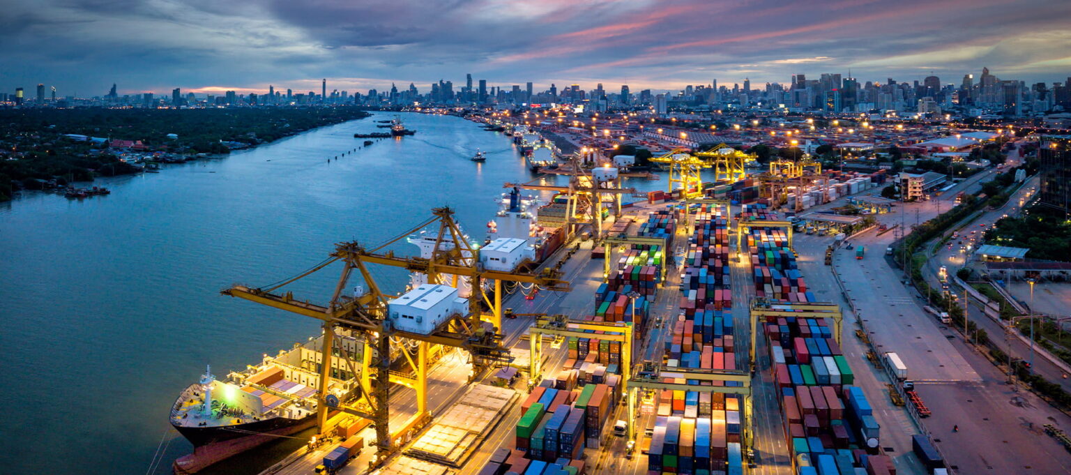 Aerial view of a busy shipping port at sunset, with cranes loading cargo containers onto ships and a city skyline in the background.