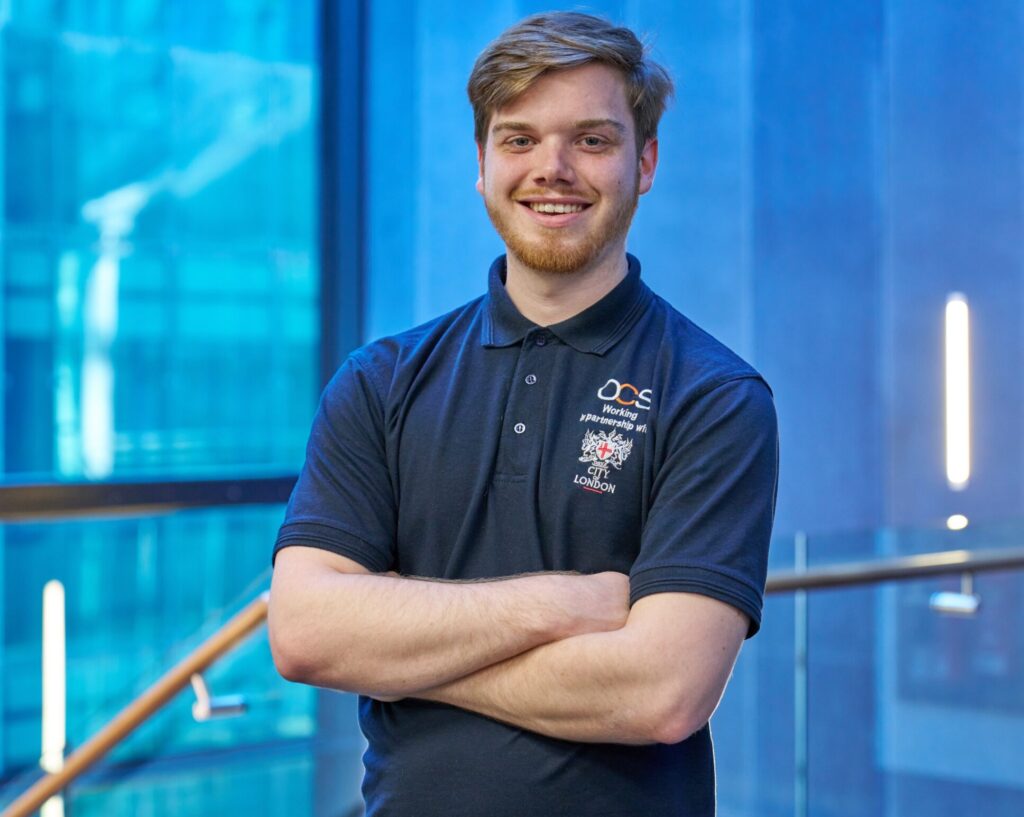 A smiling young man with short hair and a beard stands indoors with arms crossed, wearing a black polo shirt with embroidered logos. The background features glass walls and blue lighting.