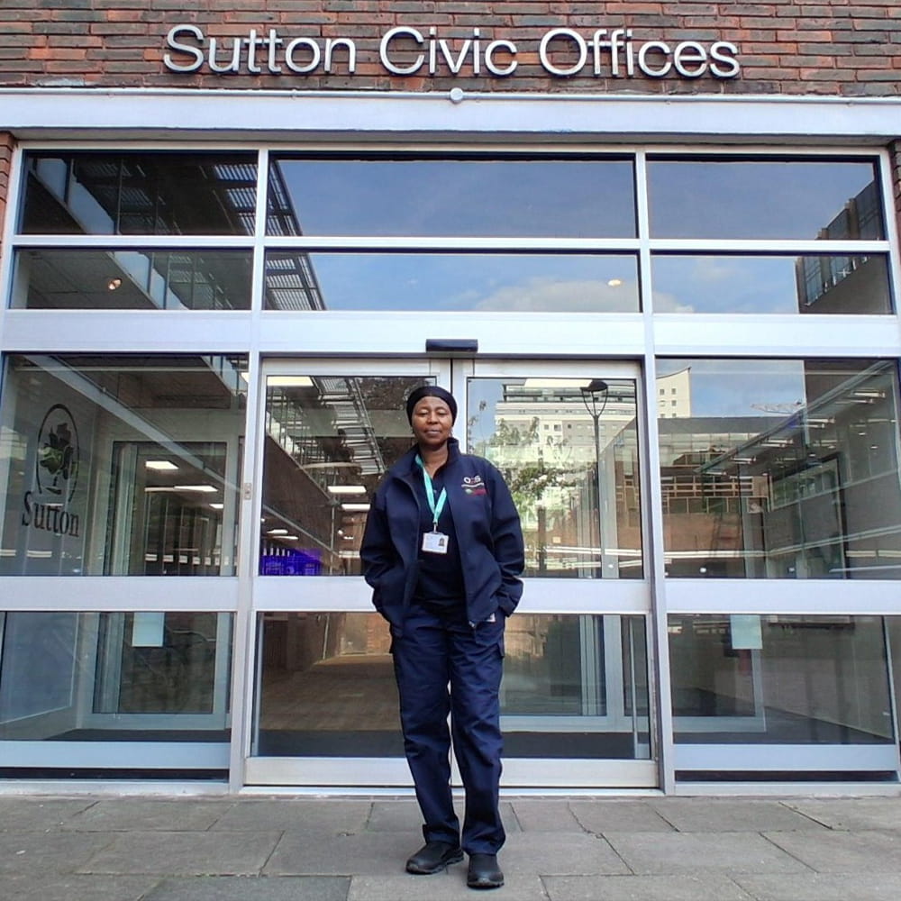 A person in a dark uniform stands in front of the glass entrance to Sutton Civic Offices, with the sign above and buildings reflected in the windows.