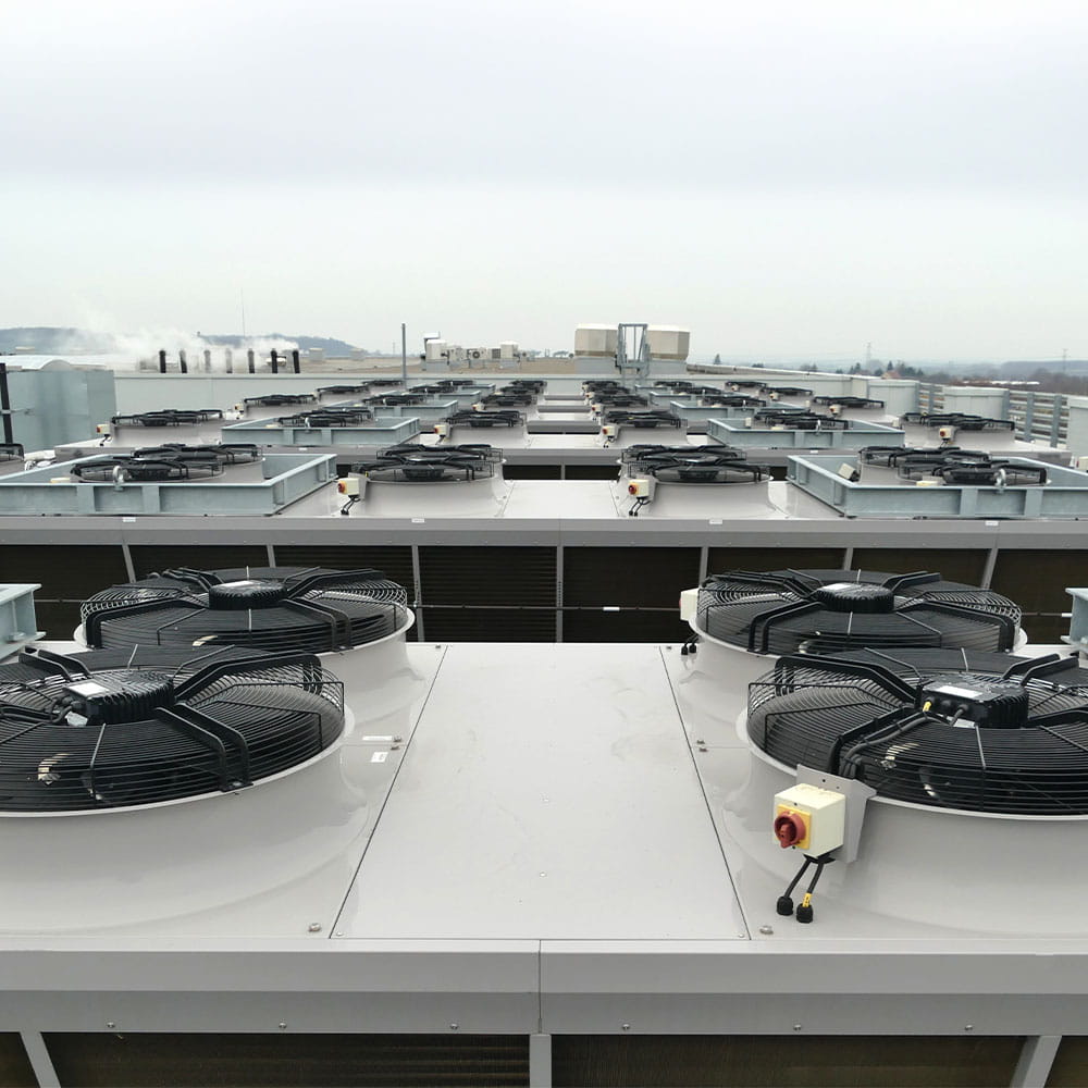 Large industrial HVAC units with multiple circular fans are lined up on a rooftop under a cloudy sky, with distant buildings and chimneys visible in the background.