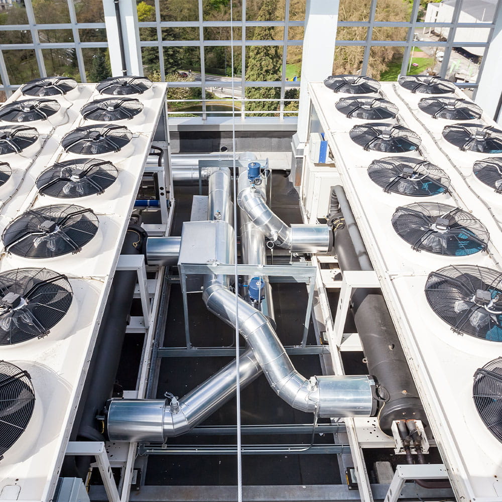 Large rooftop air-conditioning units with multiple fans and exposed silver ducting, seen from above, inside a building with large windows showing trees and a road outside.