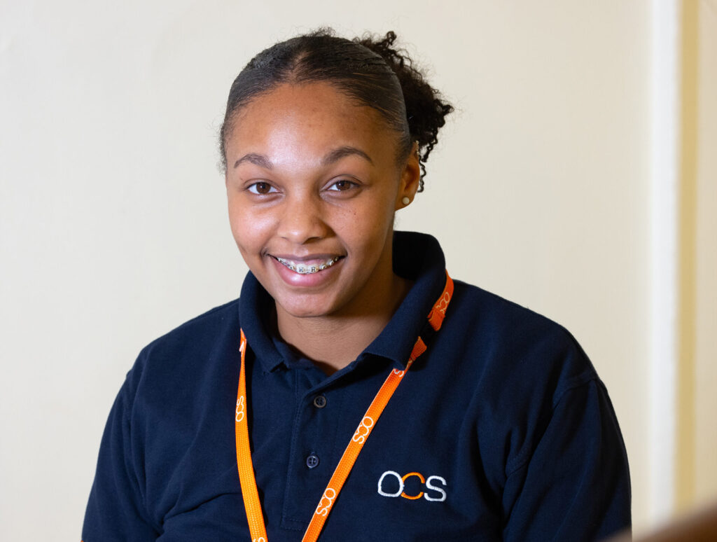 A woman wearing an OCS-branded navy shirt and orange lanyard smiles whilst standing indoors near a window and staircase.