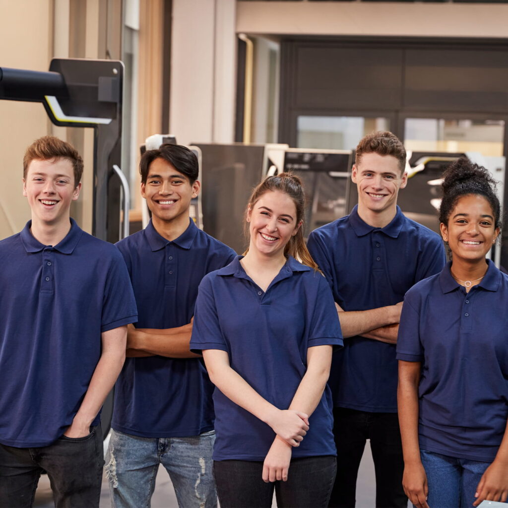 Five young adults wearing matching navy blue polo shirts stand side by side indoors, smiling and posing for the camera. The background includes modern office or industrial equipment.