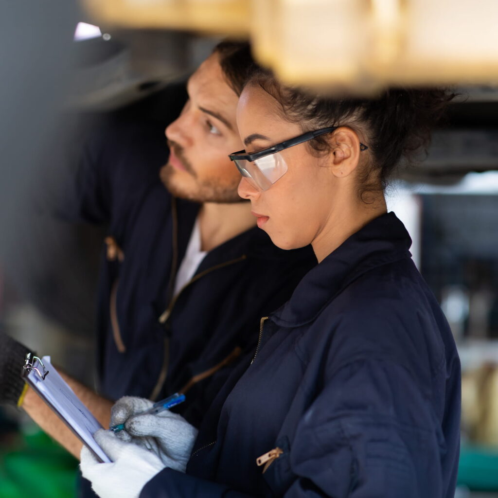 Two mechanics in overalls work together; one writes on a clipboard whilst the other inspects something off-frame. Both are focused, and the woman in front wears safety glasses.
