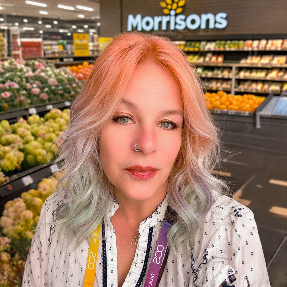 A woman with pastel pink and green hair stands in a supermarket produce section in front of a Morrisons sign, wearing a white shirt and a colourful lanyard. Shelves of fruit and vegetables are visible in the background.