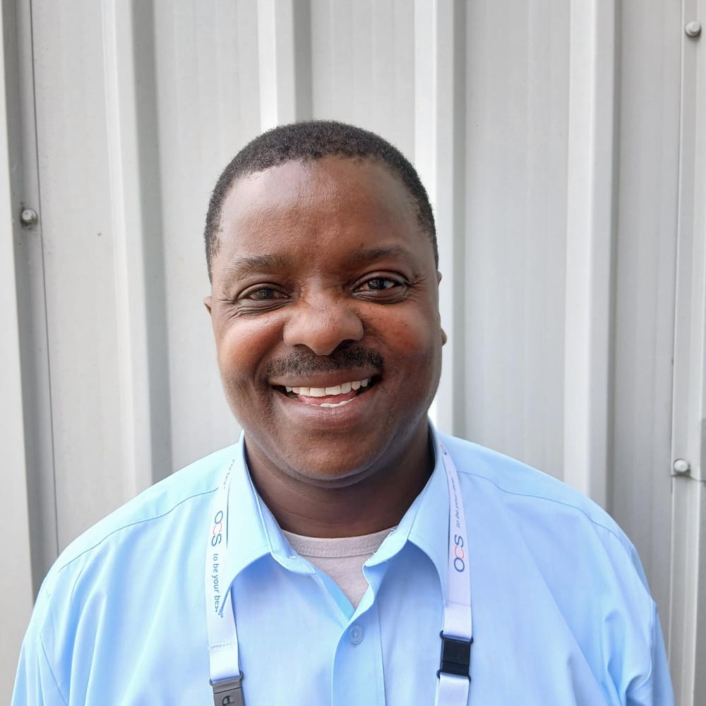 A man wearing a light blue shirt and a white lanyard smiles at the camera whilst standing in front of a grey corrugated metal wall.