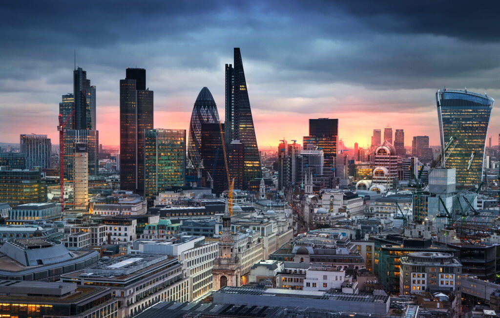 A panoramic view of London’s city skyline at sunset, featuring modern skyscrapers like The Gherkin, The Walkie-Talkie, and the Leadenhall Building among historic and older buildings under a dramatic, cloudy sky.