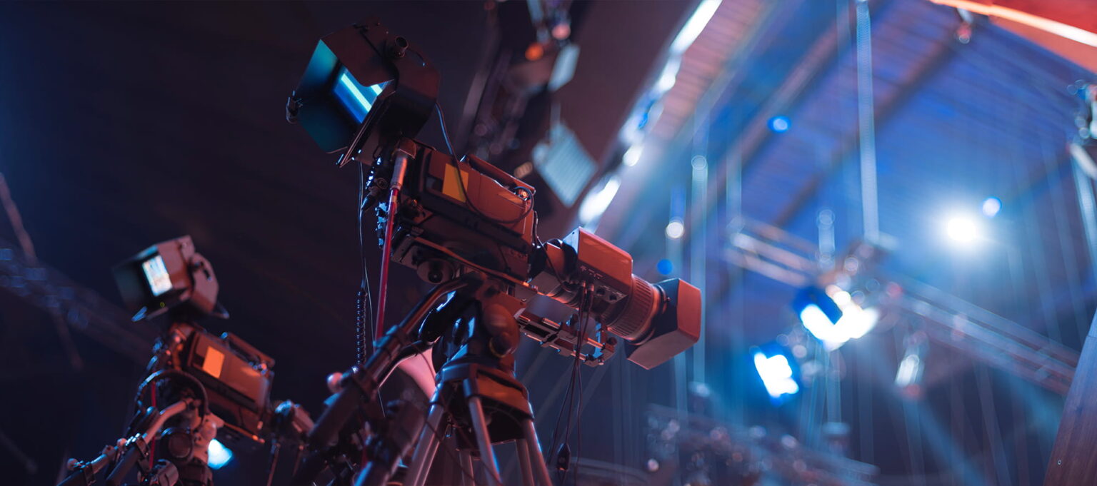 Several professional video cameras on tripods are set up in a studio with bright lights and rigging visible in the background, suggesting a television or film production environment.