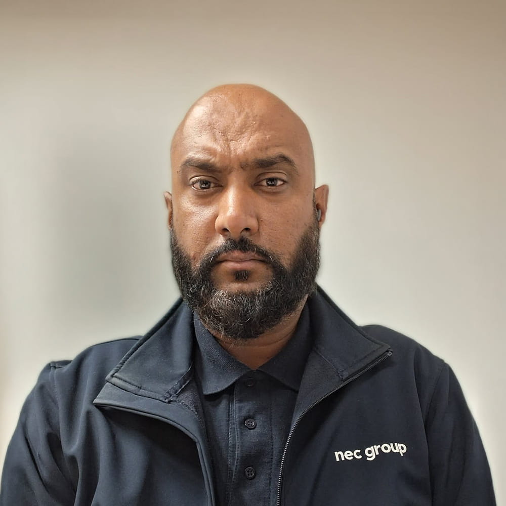 A man with a beard wearing a dark jacket with NEC Group logo, standing against a plain light background, looking directly at the camera with a neutral expression.