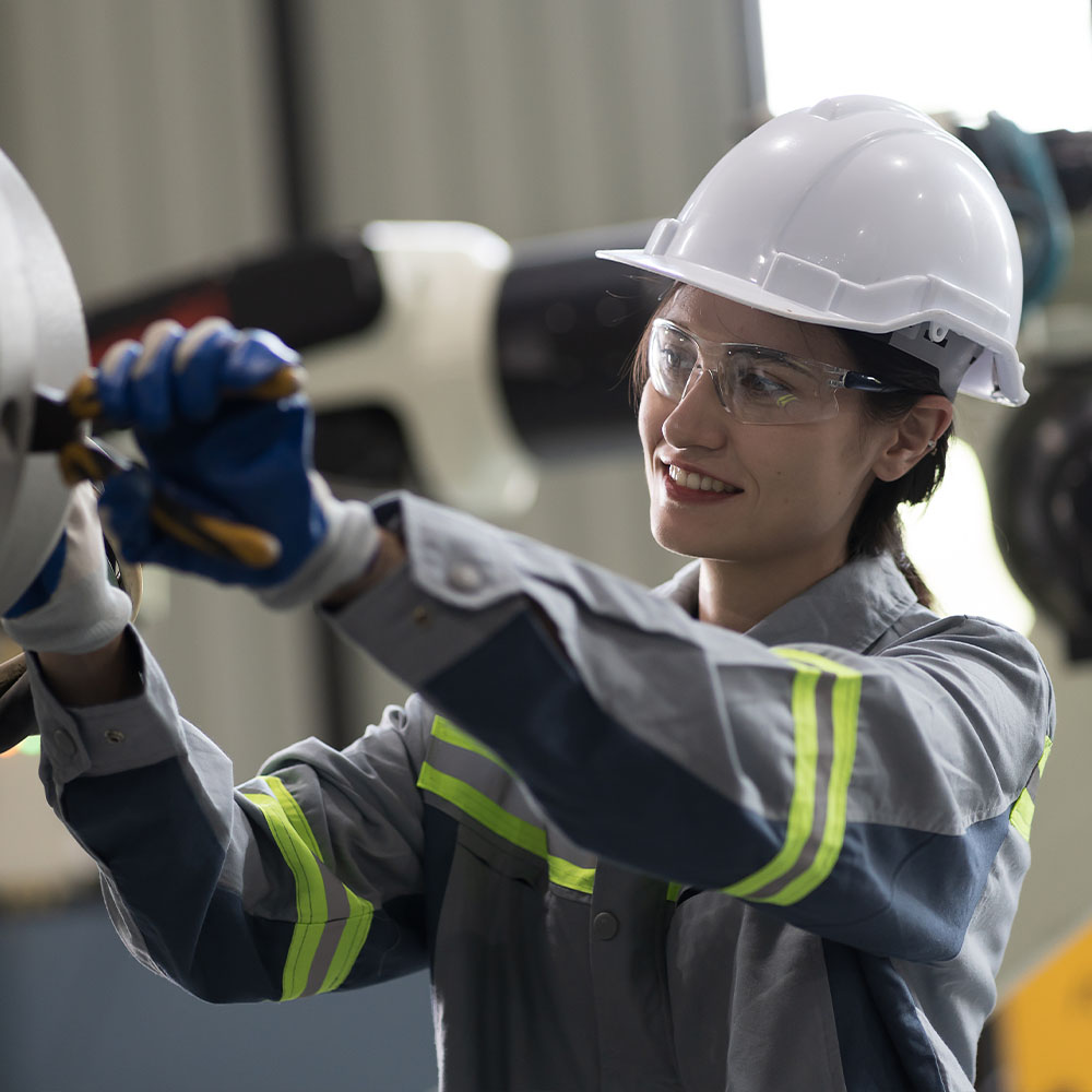 A woman wearing a hard hat, safety glasses, gloves, and a high-visibility work jacket is using a tool to adjust machinery in an industrial setting.