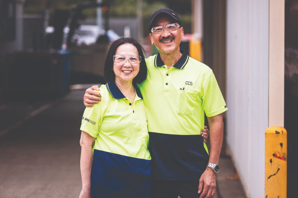 Two people wearing yellow and blue work uniforms stand side by side, smiling at the camera. One has their arm around the other's shoulder. They appear to be outside near a building.