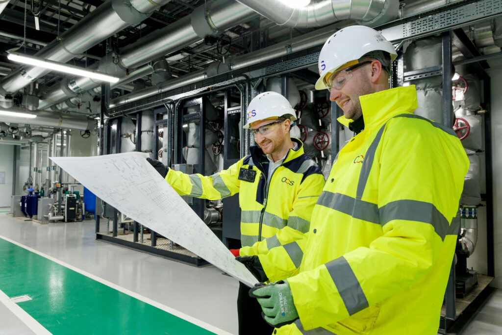 Two engineers wearing yellow high-visibility jackets and white helmets are reviewing blueprints inside an industrial facility with metal pipes and machinery in the background.