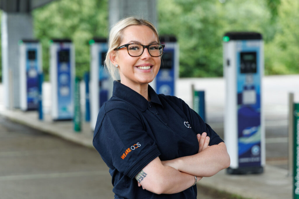 A woman with blonde hair and glasses smiles with arms folded, wearing a dark blue polo shirt. She stands in front of electric vehicle charging points outdoors.