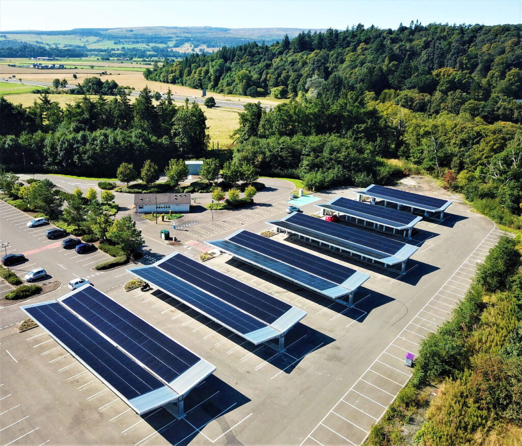 A large car park with several rows of solar panel canopies, surrounded by trees and fields. Only a few cars are parked, and green hills are visible in the background under a clear sky.