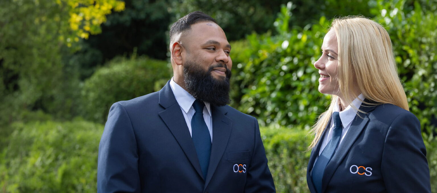 Two people in matching navy blue suits with OCS logos stand outside, smiling at each other, with green bushes and trees in the background.