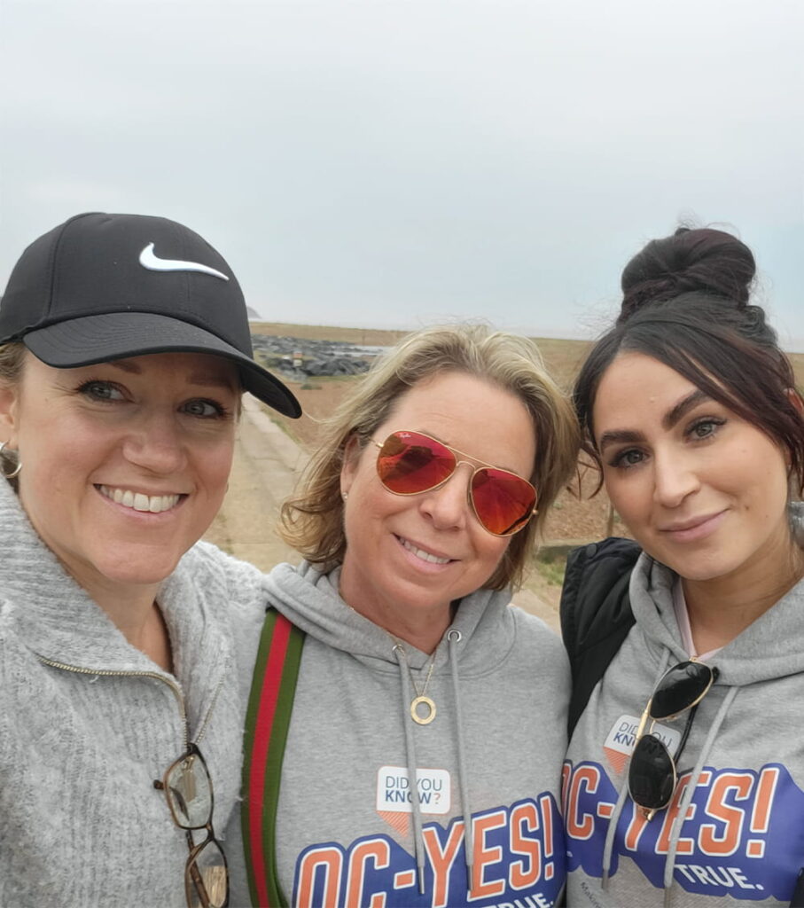 Three women smiling outdoors on a cloudy day. Two wear grey “OC-YES!” hoodies, one with sunglasses. One wears a black Nike cap. A dirt path and open landscape are in the background.