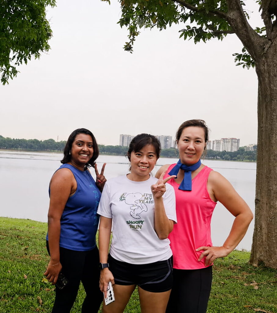 Three women posing and smiling outdoors near a lake, with trees and buildings in the background. One woman wears a blue top, another a white shirt, and the third a pink top. All are making peace signs with their fingers.