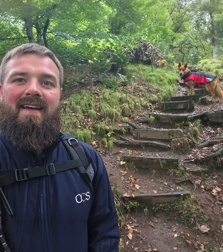 A man with a beard smiles while taking a selfie on a forest trail; behind him, a dog in a red jacket stands on wooden steps surrounded by green foliage.