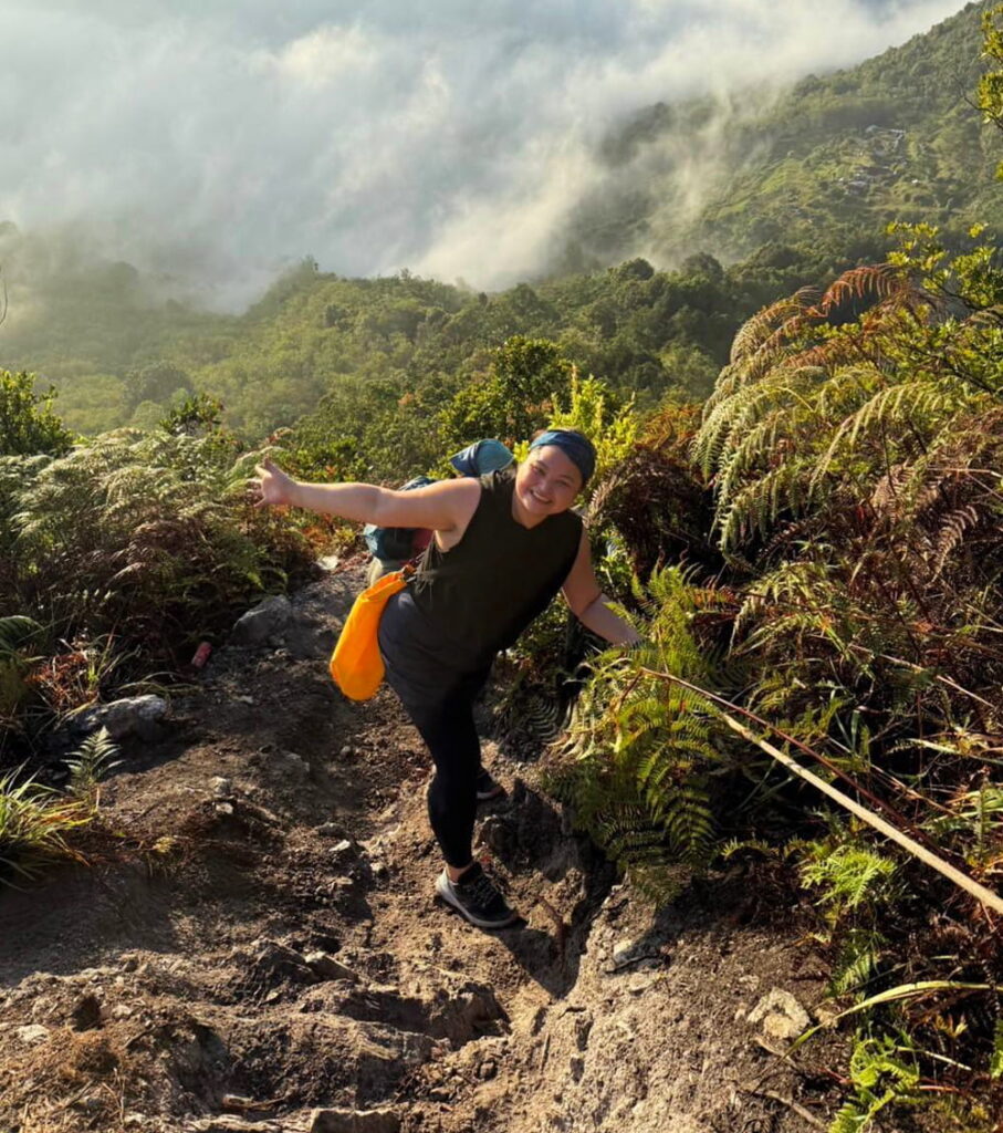 Person smiling while hiking up a rocky, steep trail surrounded by lush greenery and ferns, holding onto a rope for support, with misty mountains in the background.