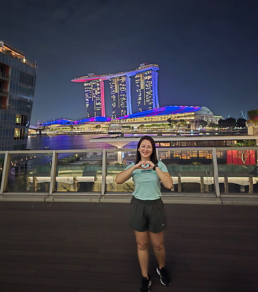 A woman stands on a boardwalk at night, making a heart gesture with her hands. Behind her, Marina Bay Sands in Singapore is brightly lit with colourful lights.