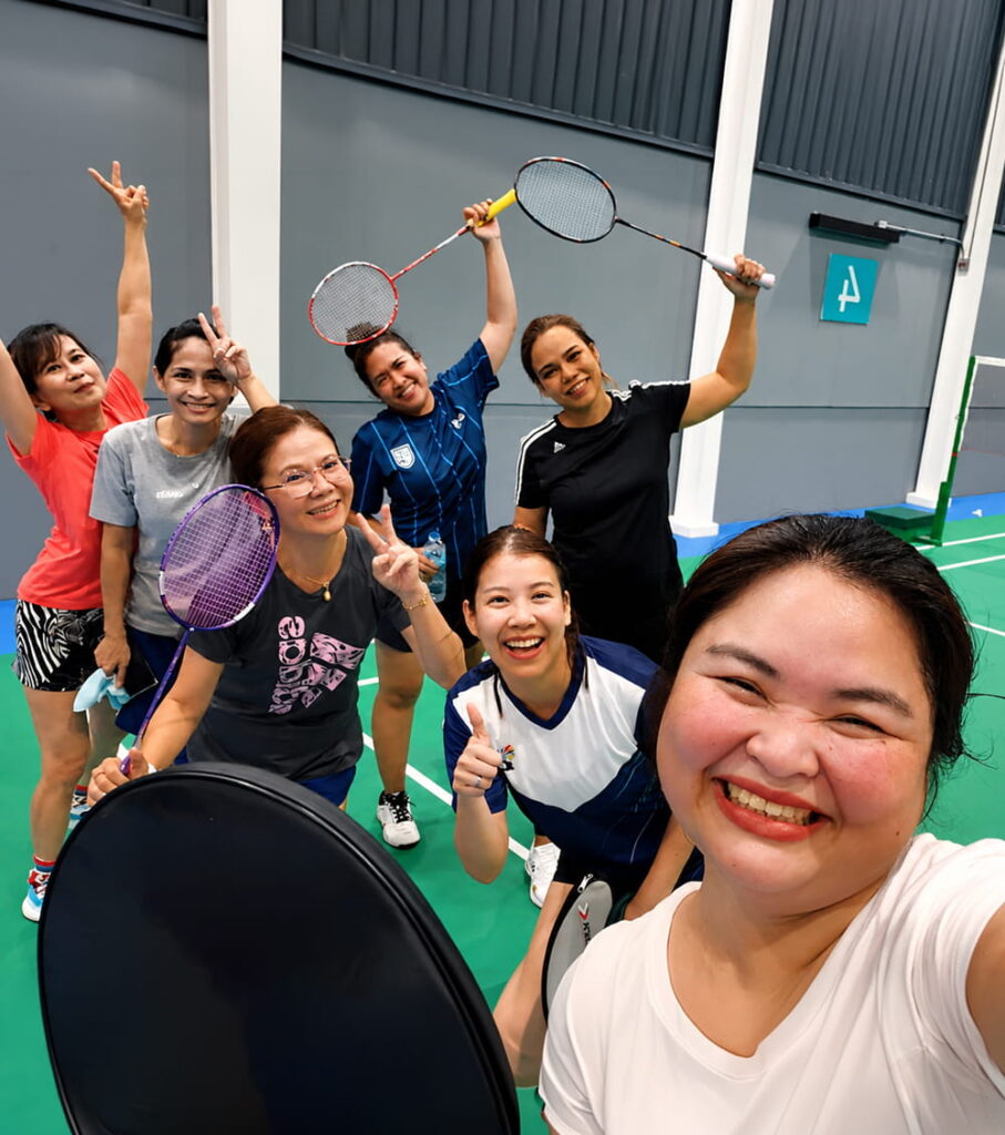 Seven women smiling and posing with badminton racquets on an indoor court, some making peace signs, all appearing happy and energetic.