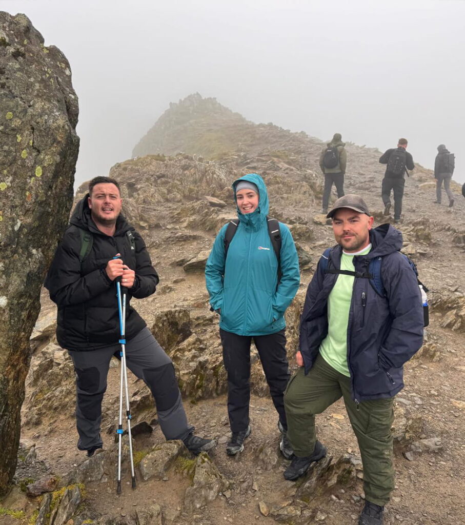 Three people in outdoor jackets stand on a rocky, foggy mountain path. Other walkers are visible in the background. The scene looks cold and misty, with rough terrain and a ridge fading into the fog behind them.