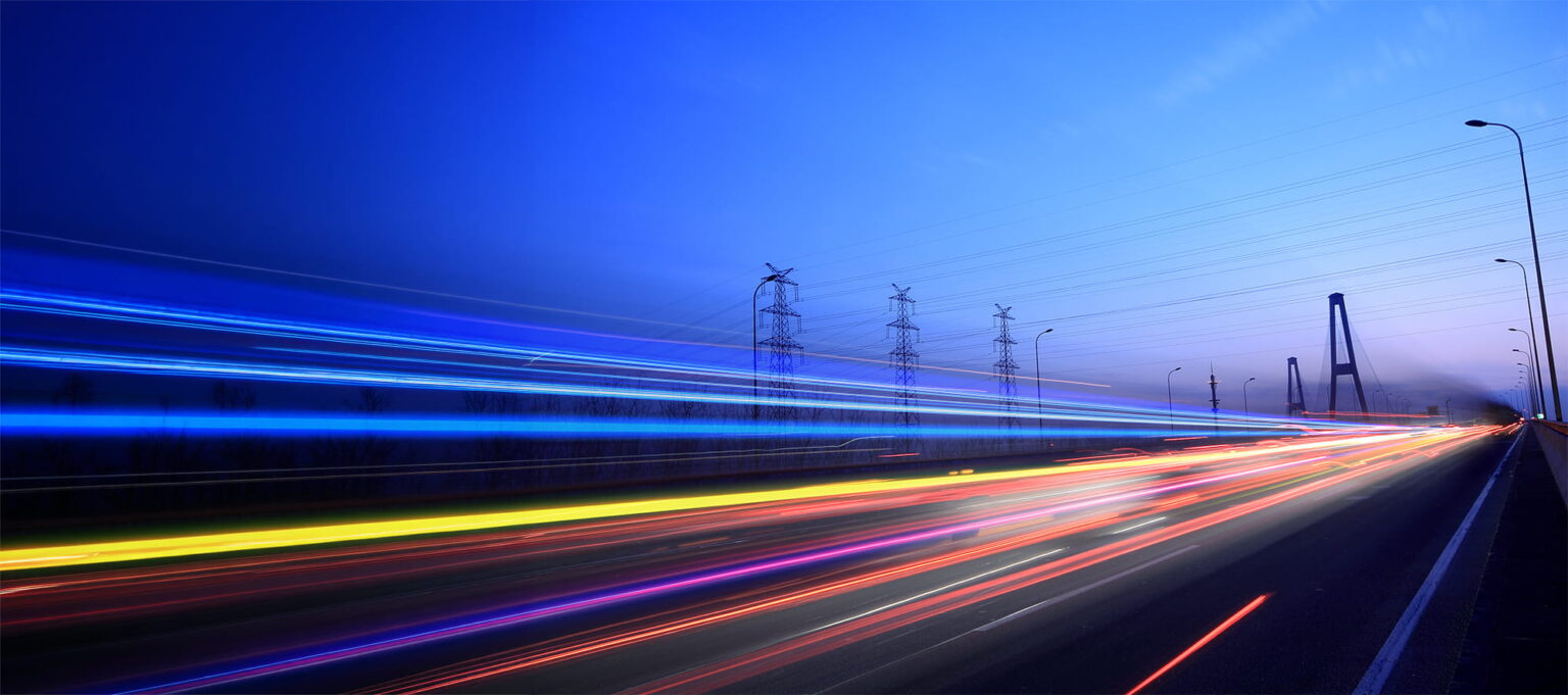 Long-exposure photo of a motorway at dusk, showing colourful light trails from moving vehicles. Power lines and pylons are on the left, with a bridge visible in the distance under a blue sky.