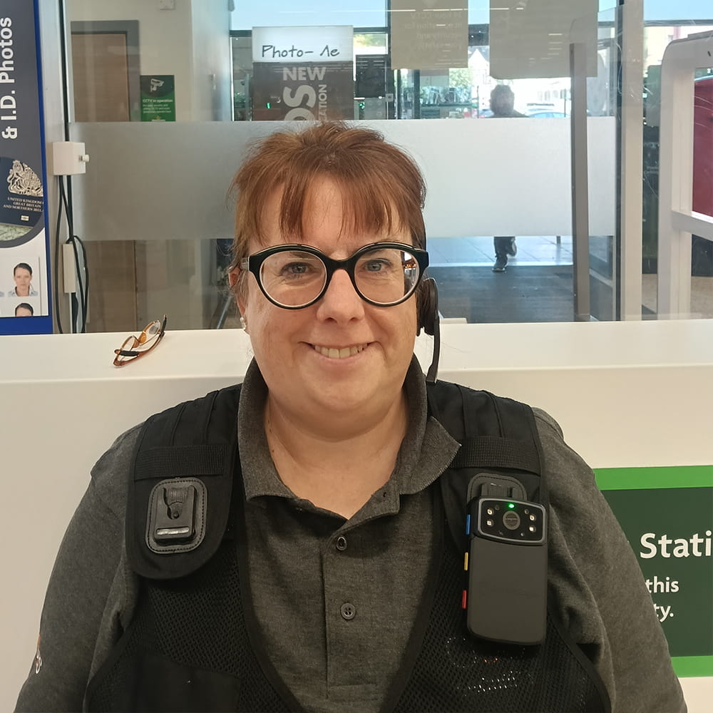 A smiling security guard with glasses stands indoors, wearing a uniform with a body camera and earpiece. Glasses rest on the counter beside her.