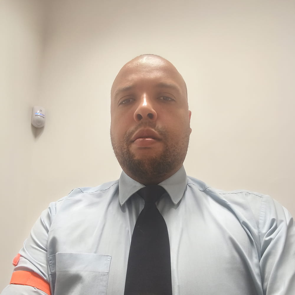 A man wearing a light blue shirt and dark tie looking at the camera in a room with plain light-coloured walls.