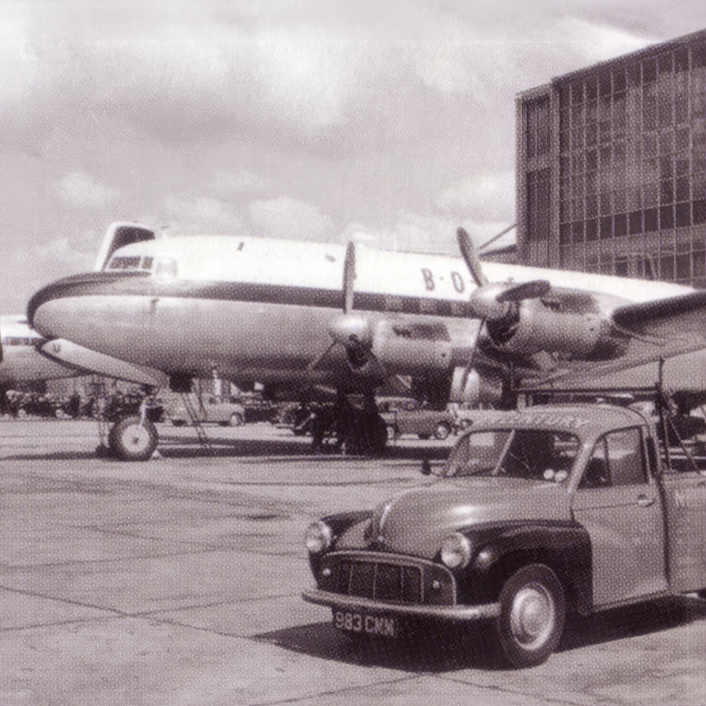 A vintage black-and-white photo shows a classic car parked in front of a large propeller aeroplane on an airport apron, with a hangar and cloudy sky in the background.