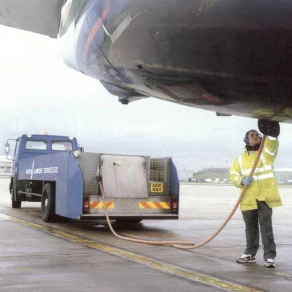 A worker in a yellow jacket refuels an aeroplane on a wet airport apron using a hose connected to a blue fuelling lorry.