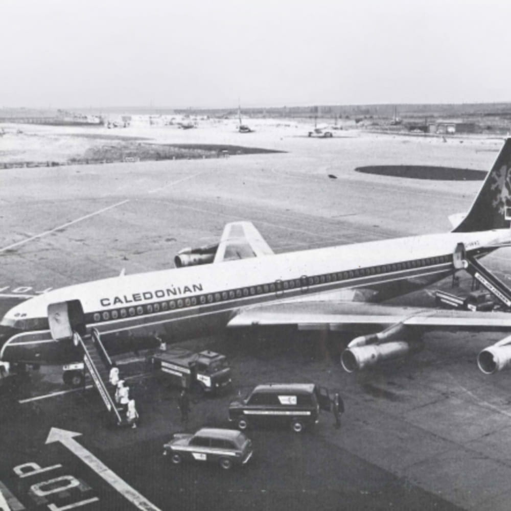 Black-and-white photo of a Caledonian aeroplane at an airport gate, with stairs leading to the door and vehicles including a van parked nearby. Distant aeroplanes and a wide apron are visible in the background.
