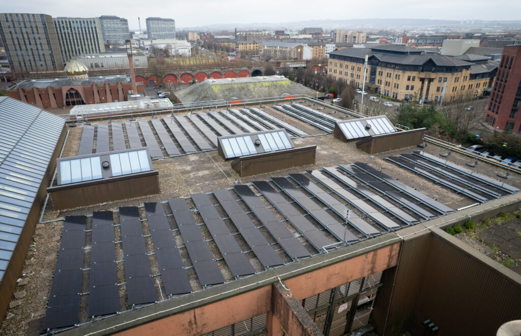 Rooftop view of a large building with multiple solar panels installed, surrounded by urban buildings, roads, and greenery on a cloudy day.