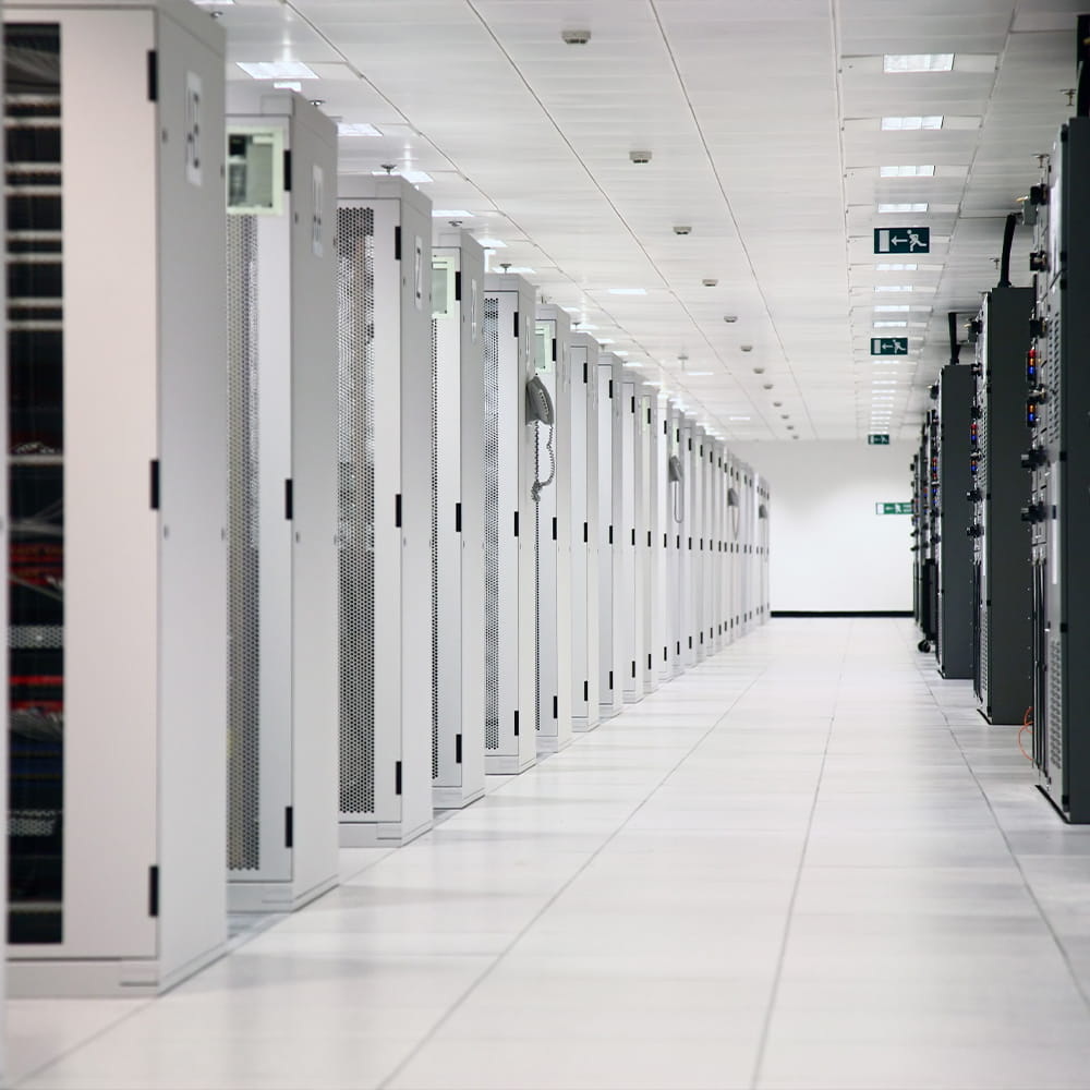 A long corridor inside a modern data centre, lined with tall, white server racks on both sides under bright ceiling lights.