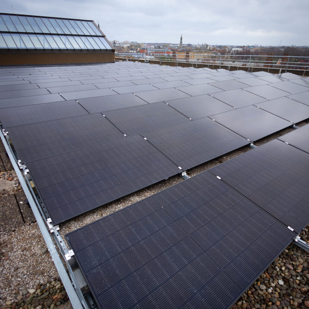 Rows of dark solar panels are installed on a flat rooftop with a cityscape visible in the distance under a cloudy sky.