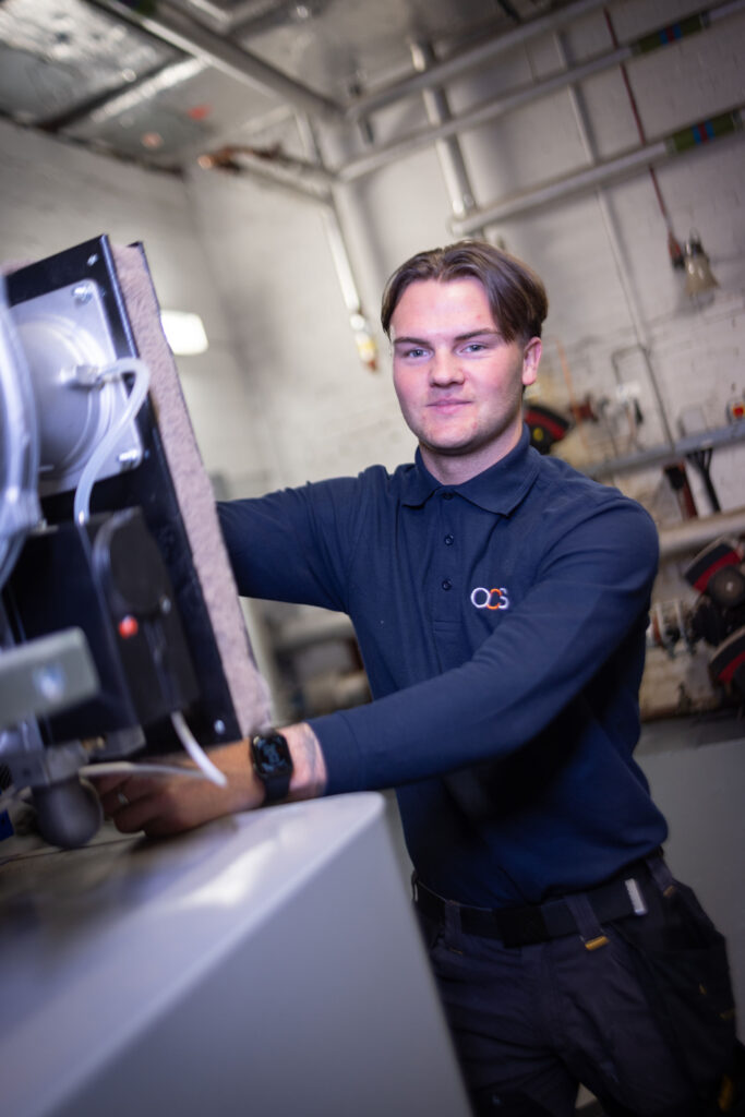 A person wearing a dark blue shirt with an OCS logo operates machinery in an industrial setting with visible pipes and equipment in the background.