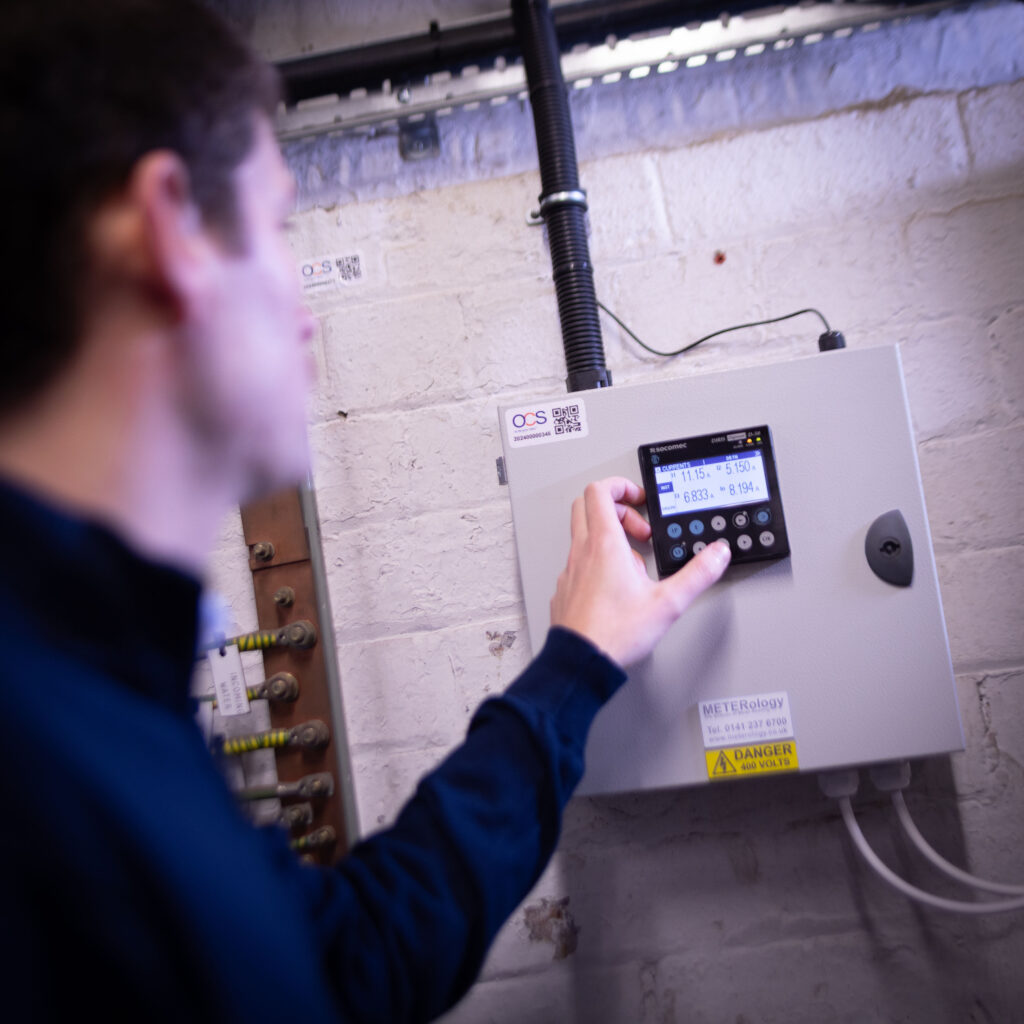A person adjusts the settings on an electrical control panel mounted on a white brick wall. The display screen shows various readings and numbers.