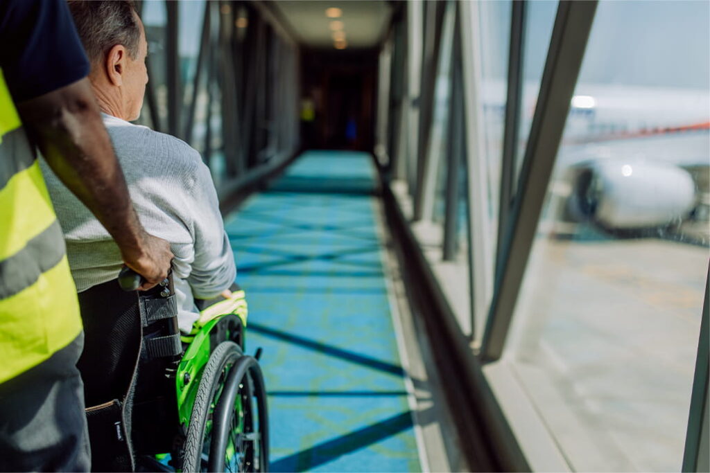 A person in a wheelchair is assisted by an airport staff member along a jet bridge towards a waiting aeroplane, seen through large windows.