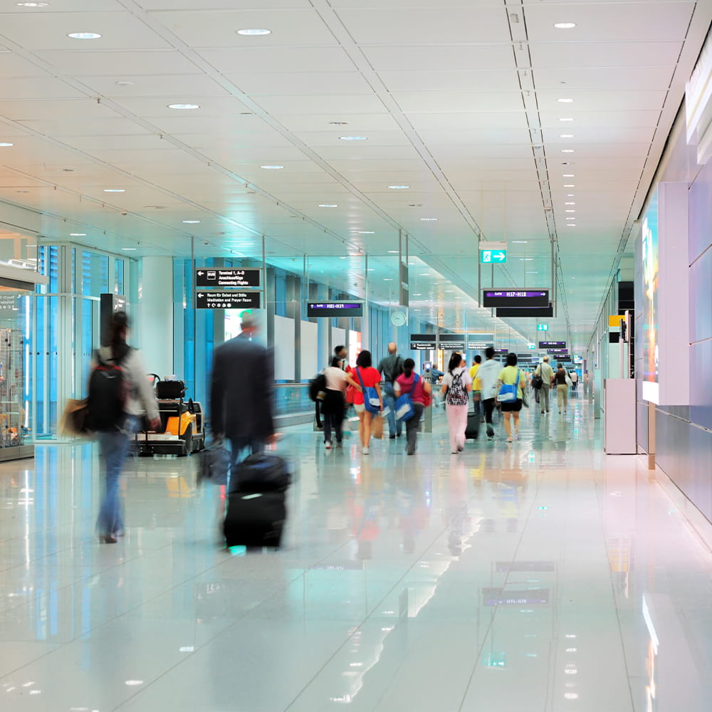 Busy airport terminal with travellers walking and pulling suitcases. Signs hang from the ceiling, and the scene is bright with reflections on the shiny floor. Some people are blurred, showing movement.
