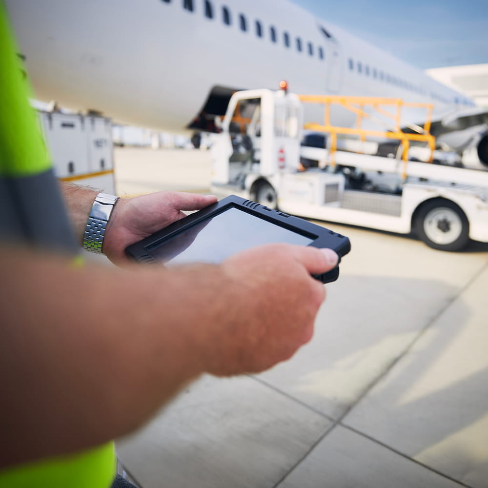 A person in a hi-vis vest uses a tablet on an airport apron, with an aeroplane and ground service vehicles in the background.