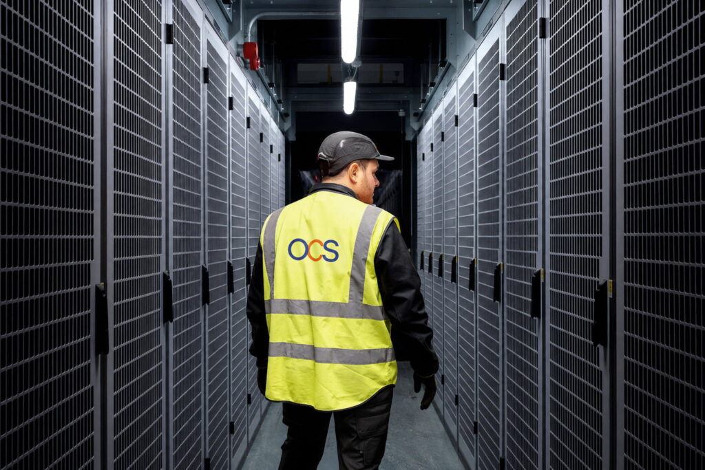 A person in a yellow OCS safety vest and black cap walks down a corridor between server racks in a data centre, illuminated by overhead lights.